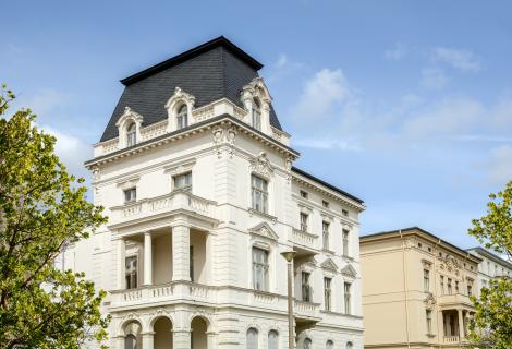 Bildrecht istock Weiße historische Stadtvilla im Gründerzeitstil mit aufwendig verzierter Fassade, Säulenbalkon und Mansarddach unter blauem Himmel, umgeben von grünen Bäumen.